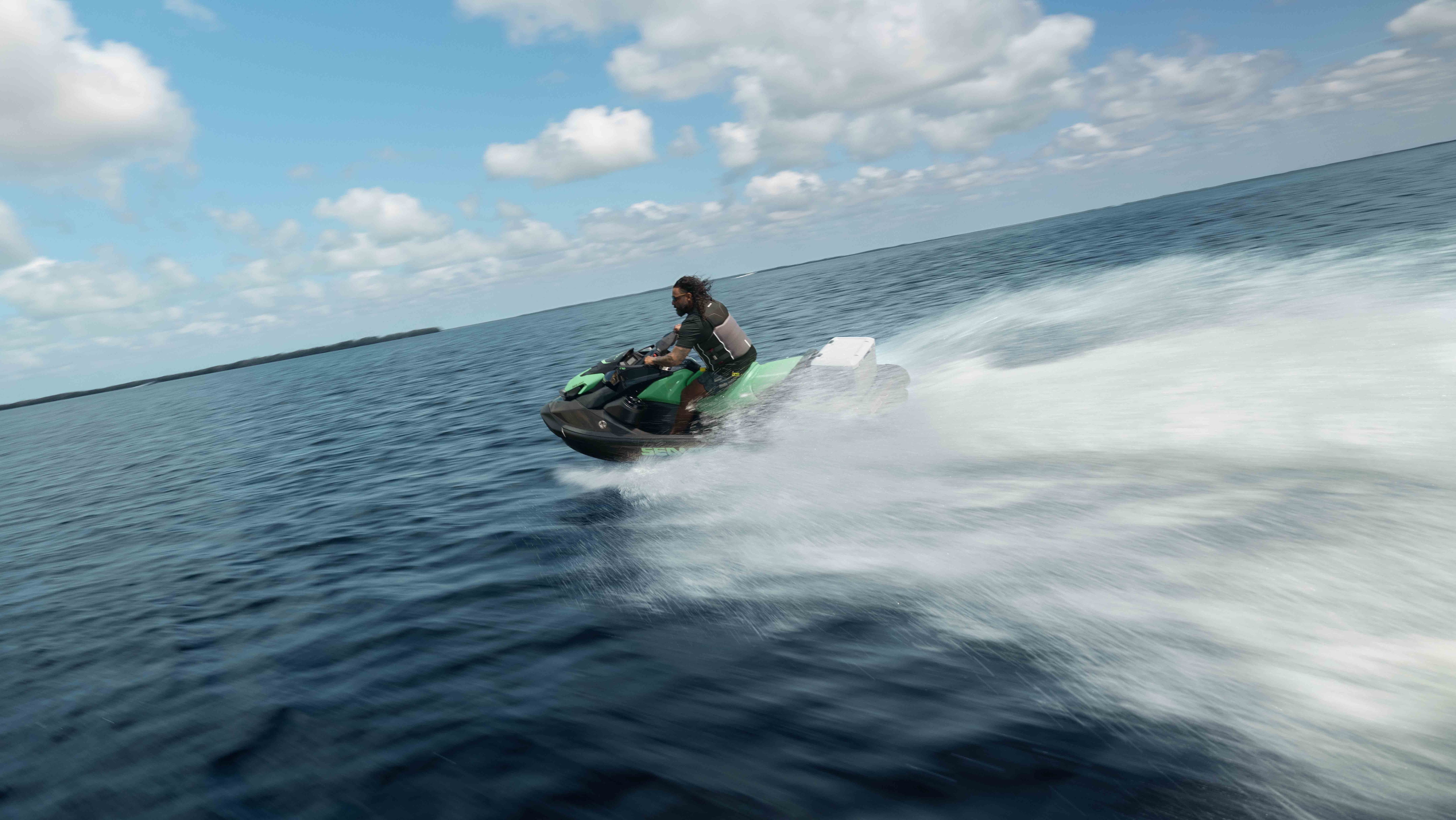 Rider on a green Sea-Doo GTI SE personal watercraft across open water, creating a large wake under a partly cloudy sky.