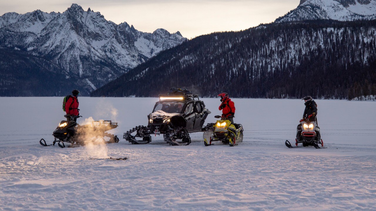 Four people on snowmobiles and an off-road vehicle gather on a snowy, frozen landscape at dusk, with mountains in the background